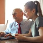 Our Team Young woman in casual clothes helping senior man in formal shirt with paying credit card in Internet using laptop while sitting at table